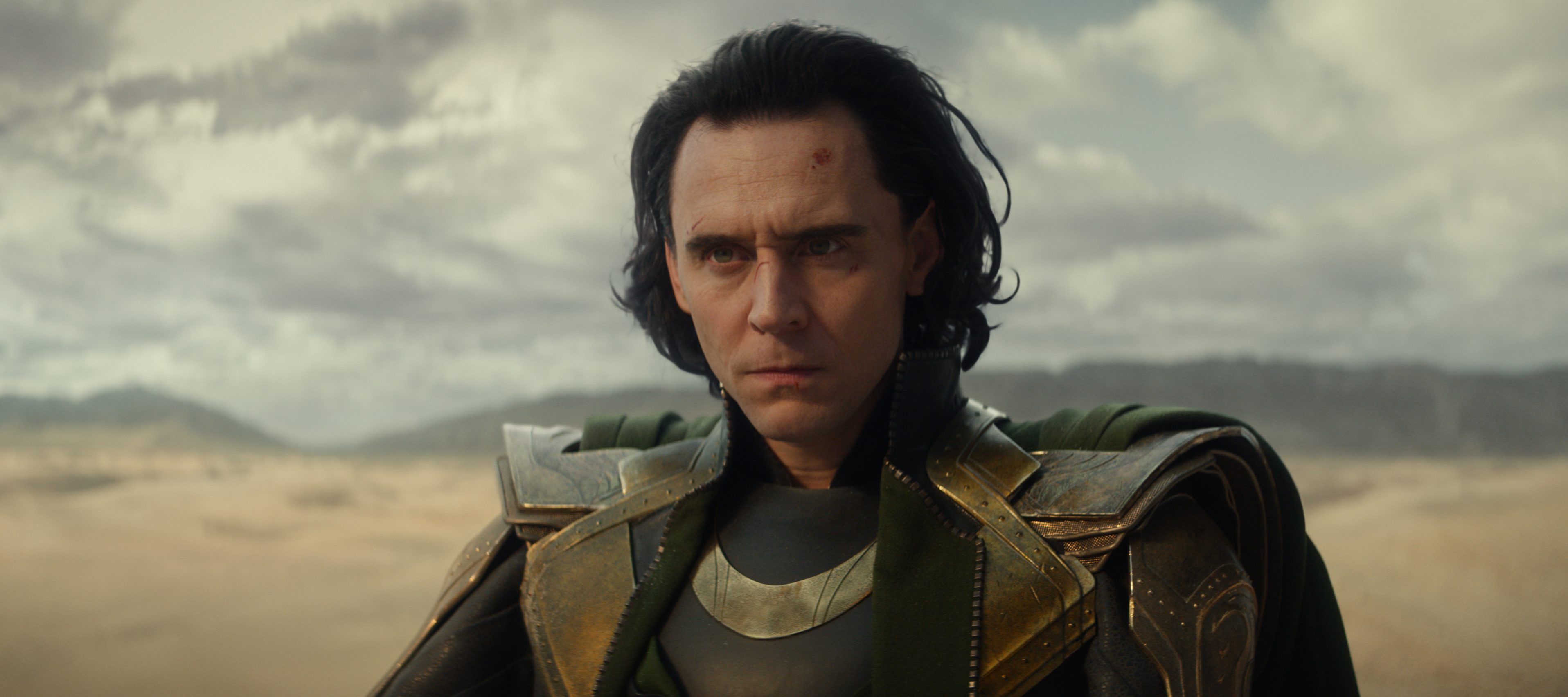 a man with long hair and a green cape looks at the camera