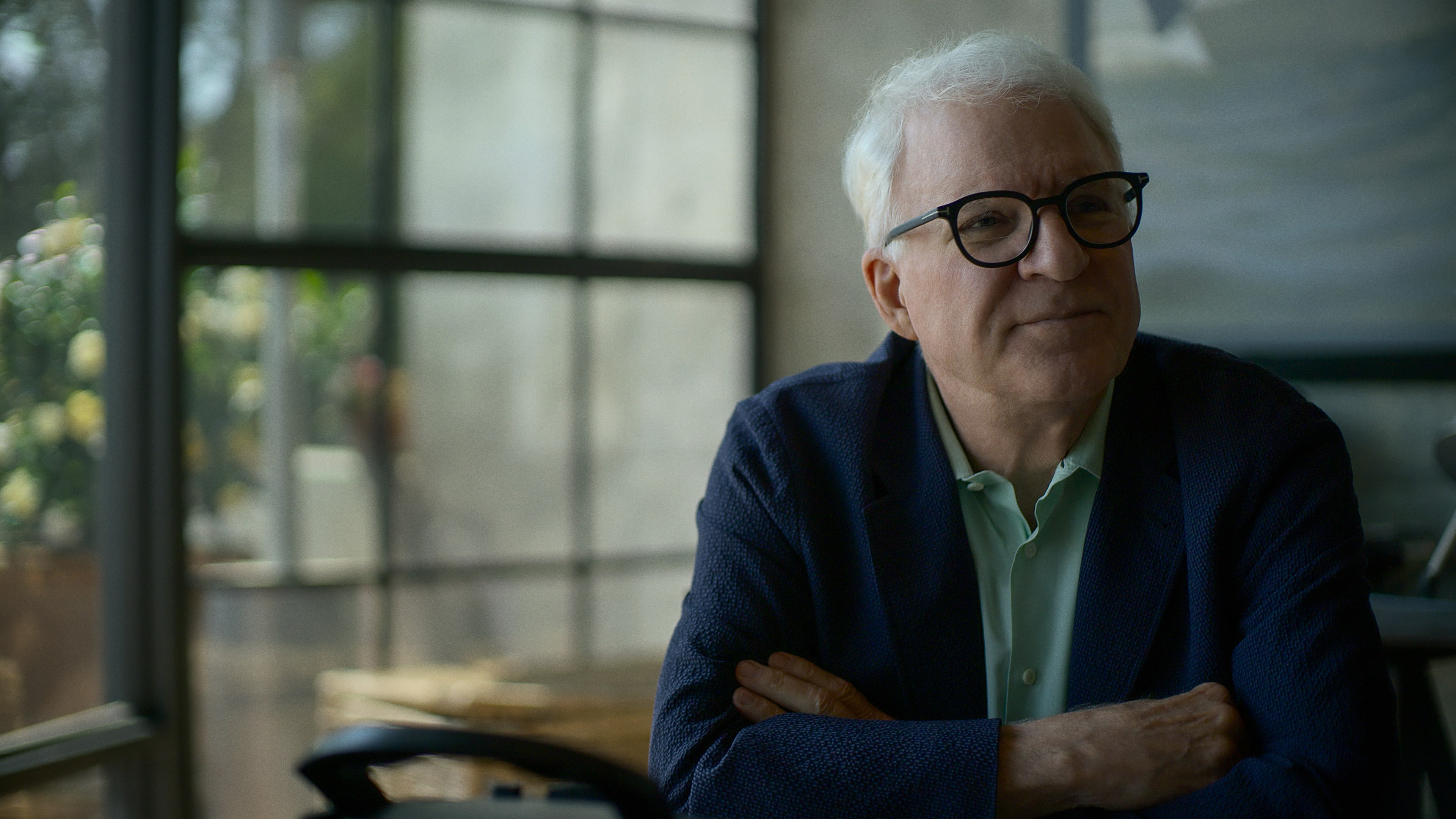 a man wearing glasses and a blue jacket sits with his arms crossed