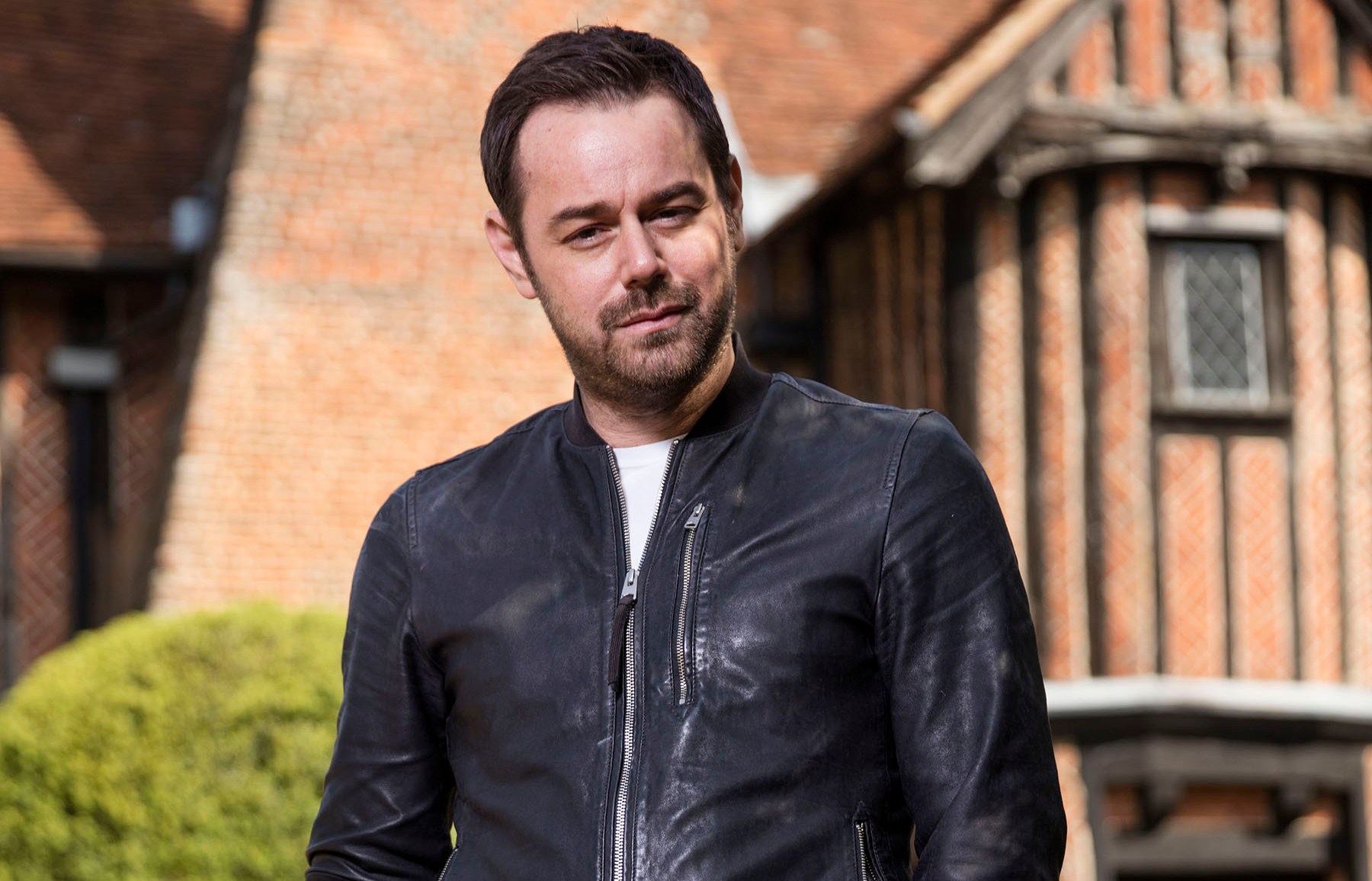 Danny Dyer in a leather jacket, holding glasses, in front of a house.