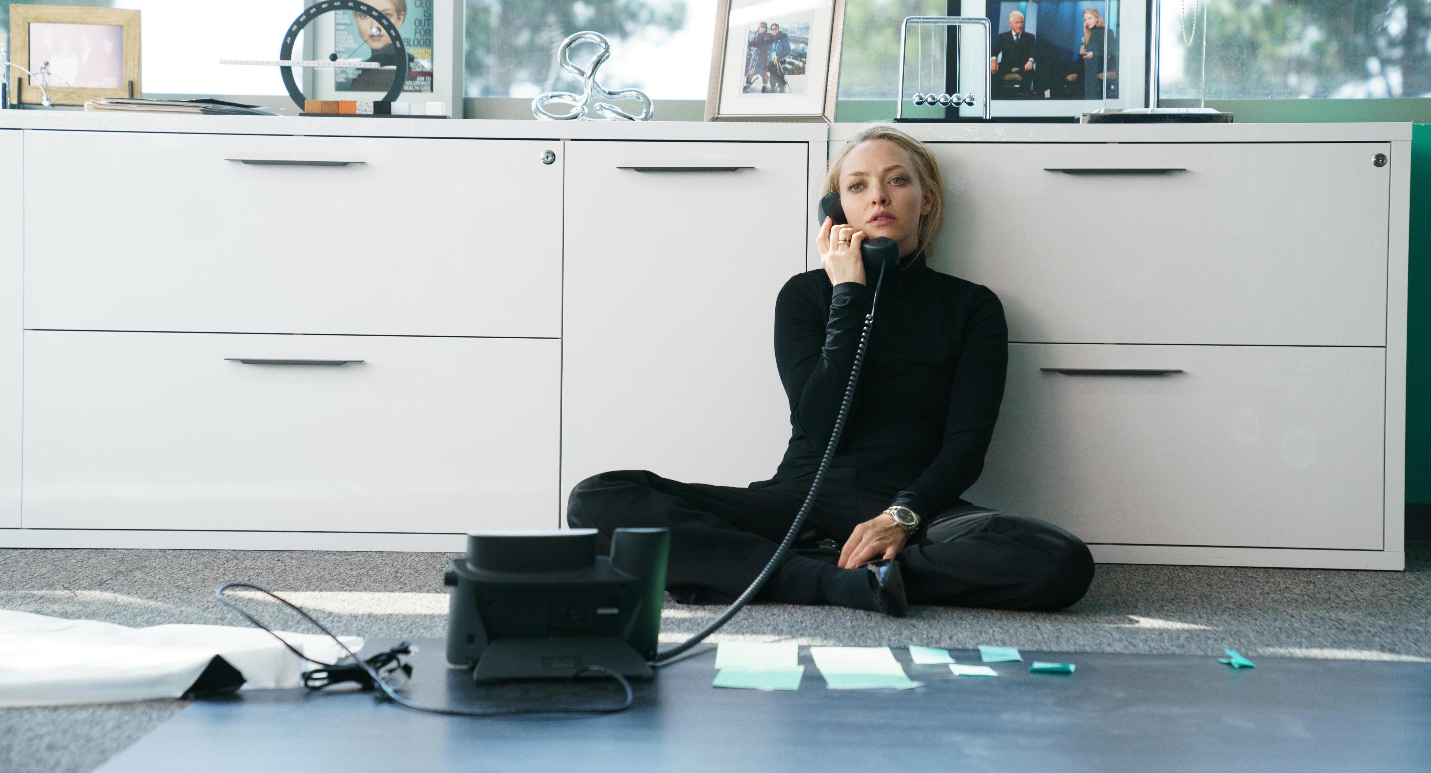 a woman sits on the floor talking on a phone with a picture of a man on the wall behind her