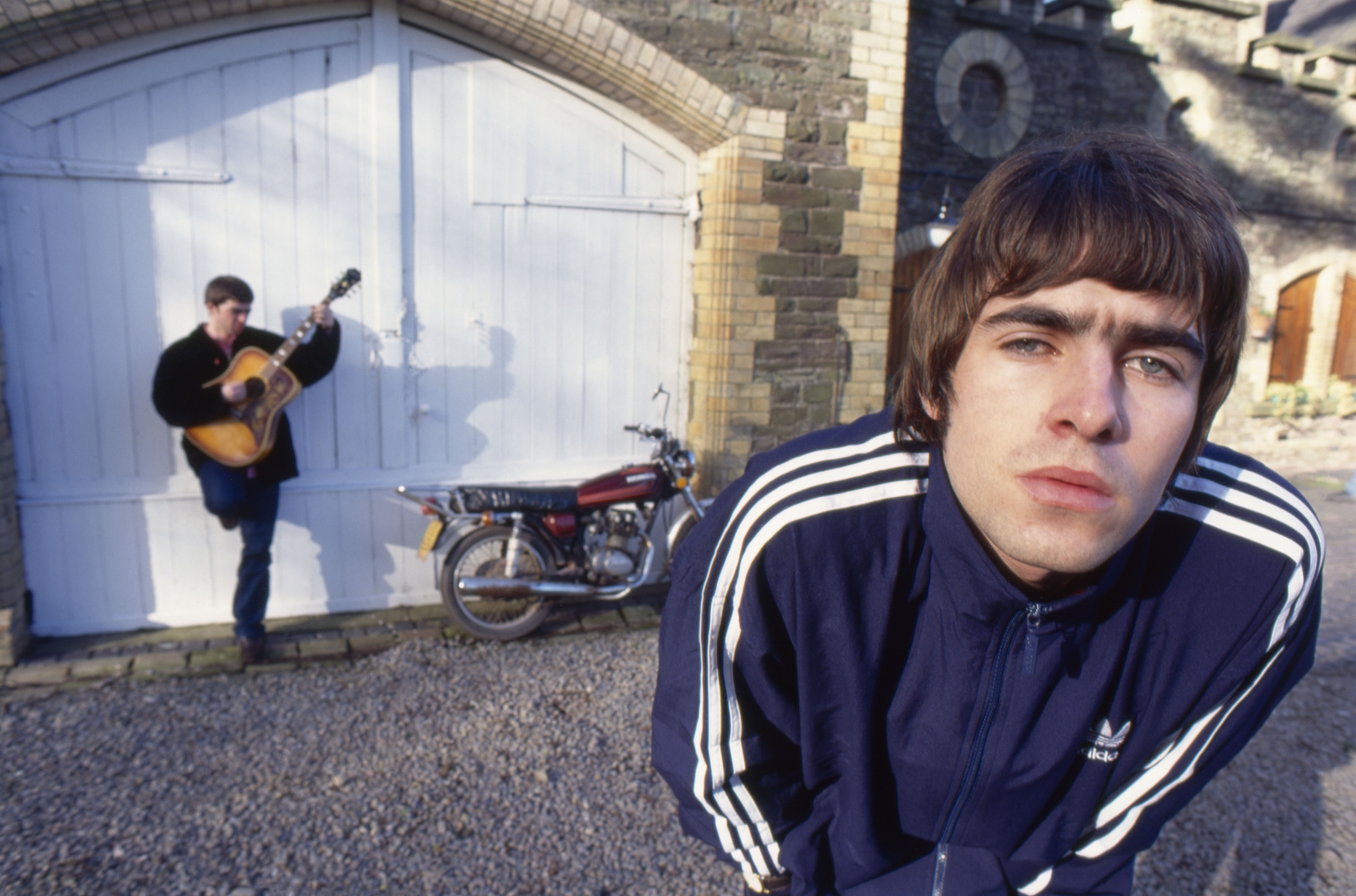 Noel Gallagher of Oasis in an Adidas tracksuit, with a bandmate playing guitar in the background.