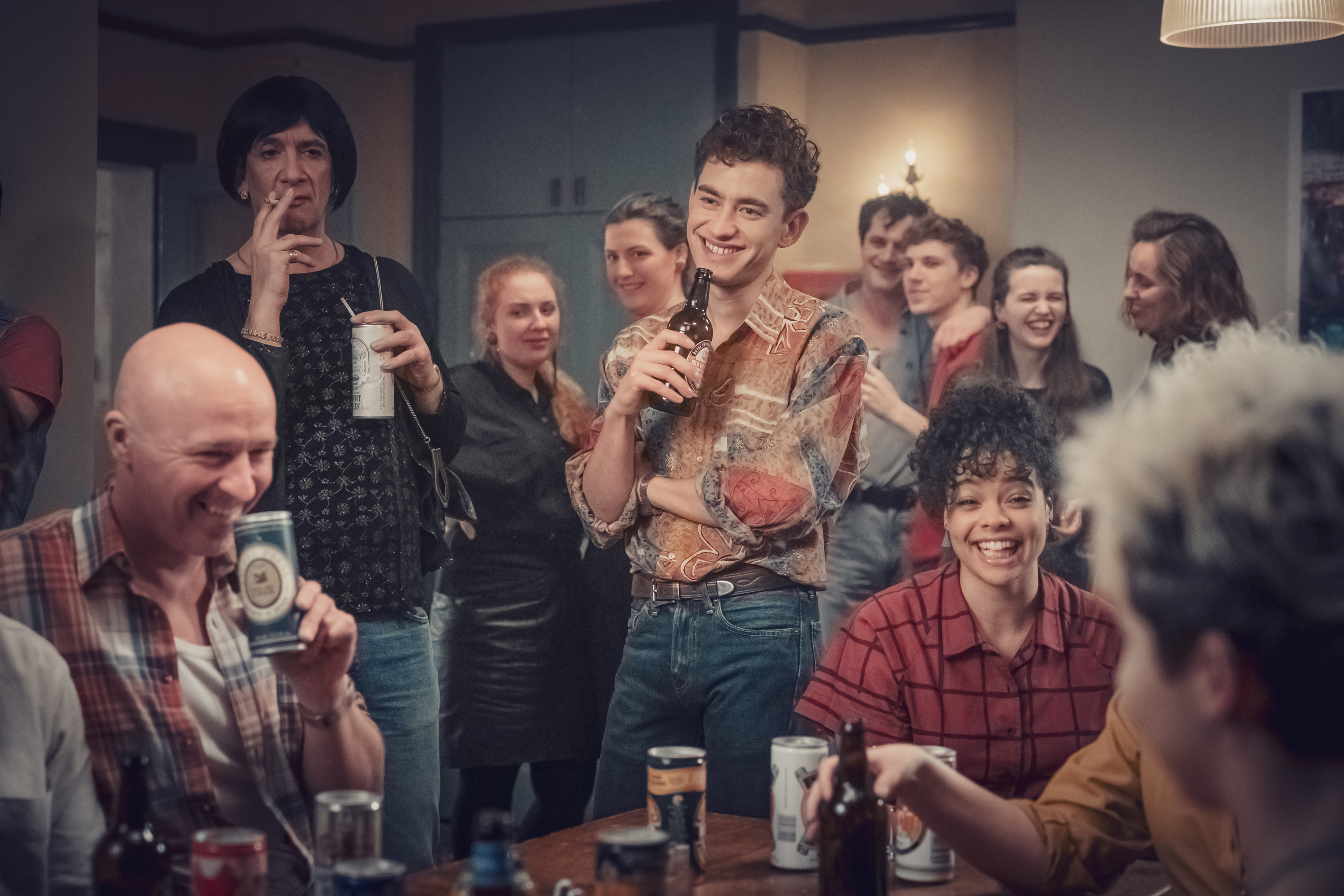 A group of people are gathered around a table with a man holding a bottle of beer.