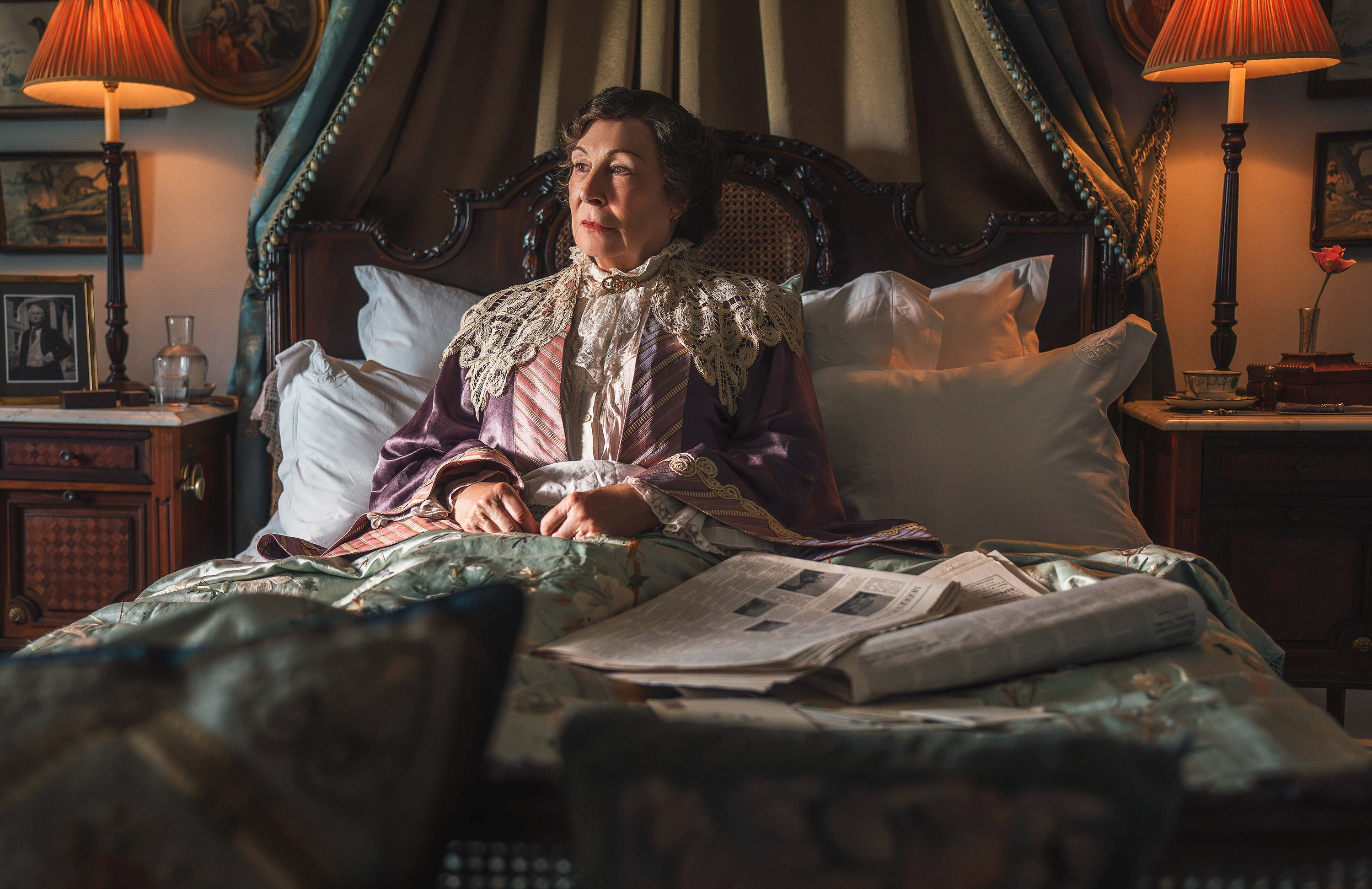 Anjelica Huston as Lady Tressilian in bed, reading newspapers.