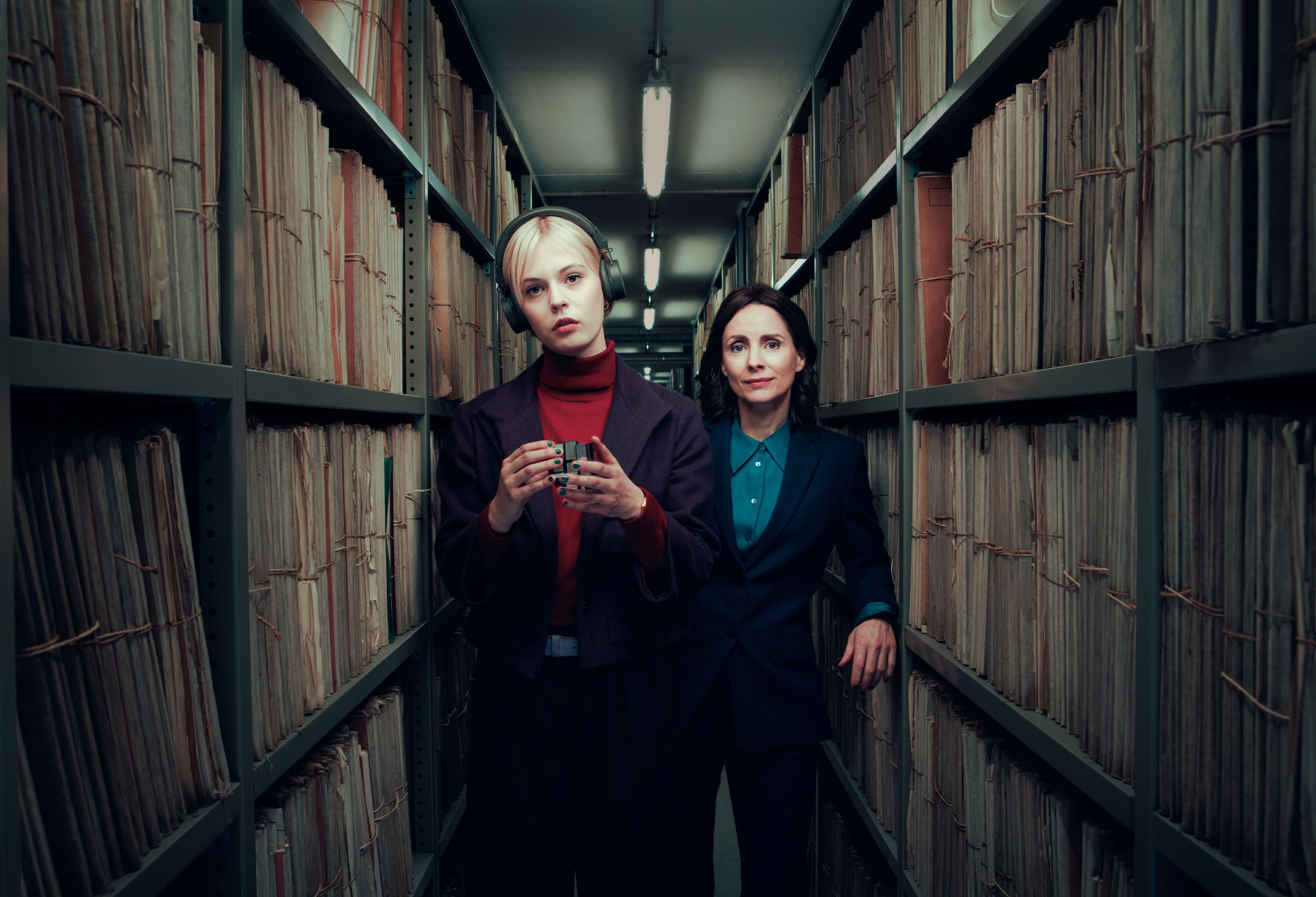 Two women stand in an archive aisle surrounded by shelves of files. One woman wears headphones and holds a Rubik's Cube.