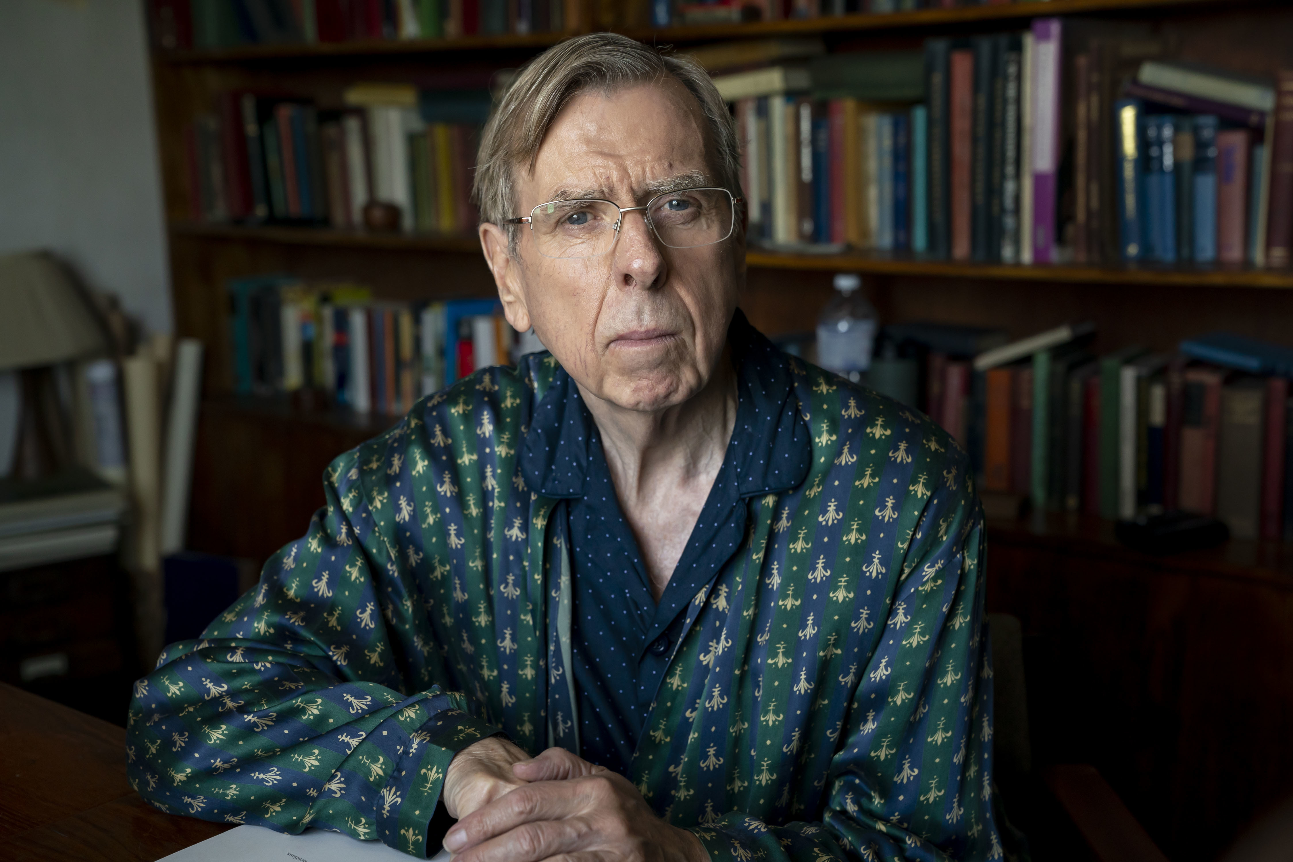 an elderly man wearing glasses and a green shirt sits in front of a bookshelf