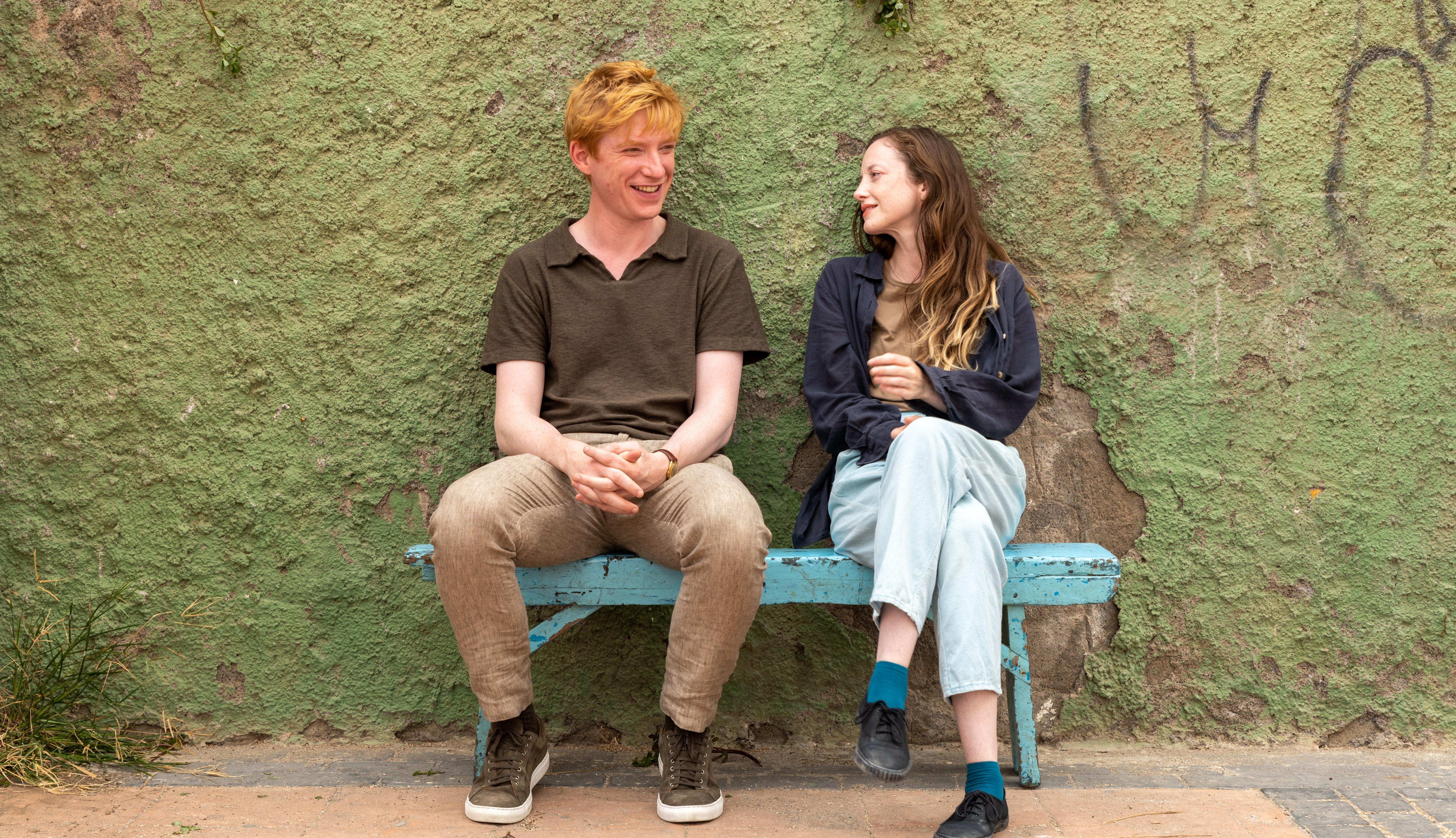 A man and a woman sit on a blue bench in front of a green wall.
